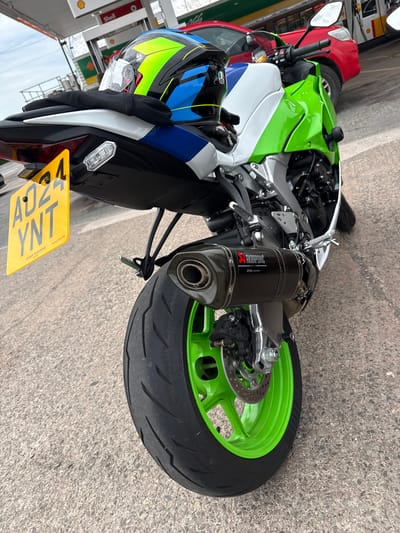 The image shows the rear view of a green and white motorcycle parked at a Shell petrol station. The motorcycle has a black seat, a black exhaust with the Akrapovič logo, and a bright yellow number plate with the registration "AD24 YNT". A helmet with green, blue, and yellow detailing rests on the rear seat. In the background, a red car is visible, along with petrol pumps and the Shell branding. The ground is paved with concrete. The scene appears to be in the city of Much Dewchurch, United Kingdom, as indicated by the presence of the Shell petrol station, which are common in such locations.