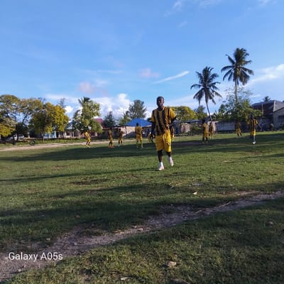 The image shows a soccer field in Nungwi, Tanzania, located at coordinates -5.726 latitude and 39.301 longitude. The field is mostly covered with green grass. There are eight men on the field, all dressed in yellow and black jerseys. They are participating in a soccer game. One man is in the foreground, walking towards the camera, while others are playing the game in the background. There are several trees in the background, including a tall palm tree, and several houses in the distance. The sky is blue with a few white clouds. The photo was taken during the daytime.