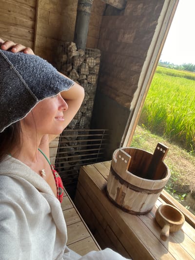A woman wearing a gray head covering and a white wrap is lying down inside a wooden structure, possibly a sauna, in Singakerta, Indonesia. The structure features a stove with stones in a cage. A wooden bucket and ladle sit on a platform nearby, with a view of lush green vegetation visible outside. The woman is lying on her back with her head tilted, and a glimpse of swimwear is visible under her wrap.