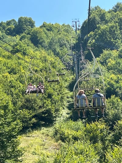 This is a telephoto shot of a ski lift ascending a heavily forested mountainside under a clear blue sky. The ski lift's chairs are visible, some occupied by people. In the foreground on the left, a chair carries three people, two adults and a child. Further up the slope, a chair carries two adults. In the distance, another ski lift tower is visible. The mountain is covered in dense green trees, with a patch of sunlit grass visible on the lower slopes. The specific location of the ski lift in Mizhhiria, Ukraine, is unknown from the image alone.
