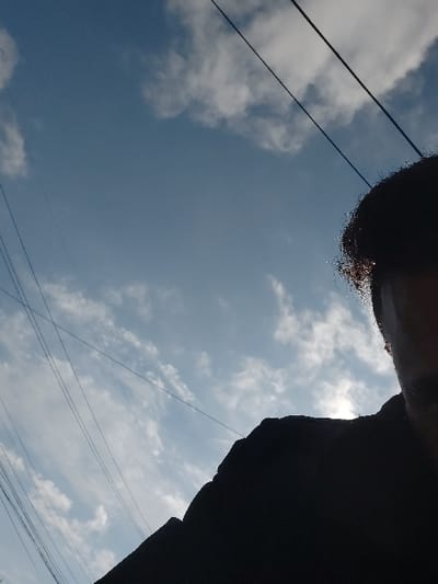 The image displays an upward-angled view of a sky scene, situated within the city of Gimpo-si, South Korea. The upper two-thirds of the frame show a clear blue sky with scattered white, wispy cumulus clouds. Several dark, thin utility lines traverse the left and upper sections of the image diagonally. The lower right quadrant is occupied by the partial, heavily silhouetted profile of an individual, photographed from a low perspective. Details visible include the top of a head with dark hair, which exhibits a bright, luminous outline caused by a strong light source, likely the sun, positioned directly behind the person. A small, indistinct portion of the individual's right ear and the side of their face are discernible in shadow, above a dark, formless mass that represents the upper body or clothing. No specific actions or interactions are evident.