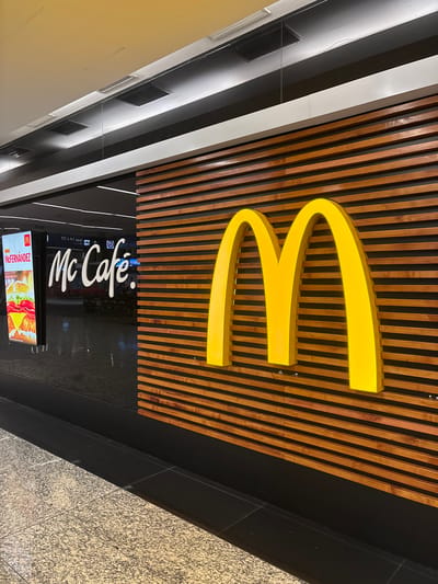 The image shows the exterior of a McCafé located in Aeropuerto Internacional Ezeiza, Argentina. The main focus is on the bright yellow McDonald's "M" logo prominently displayed on a wooden slatted wall. To the left of the logo, a large glass window reveals the interior of the establishment. Through the window, a digital advertisement for "McFernández" is visible, featuring a close-up of a burger. The McCafé logo is also visible on the glass in white lettering. The floor in front of the establishment is made of speckled grey and black tiles. The lighting appears to be artificial, likely from overhead fixtures. The scene is clean and modern, typical of a fast-food establishment in an airport. There are no people visible in the image.