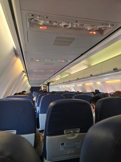 The image shows the interior of an airplane cabin. Rows of blue seats with headrests are visible, occupied by passengers. Overhead compartments line the ceiling, and light fixtures emit a warm glow. A flight attendant in a red uniform is standing near the front of the cabin, interacting with a passenger. The image appears to be taken from a passenger's seat, looking towards the front of the plane. The environment is clean and orderly, typical of an aircraft cabin. There is no information in the image that suggests the location is Ramunia Dua, Indonesia.