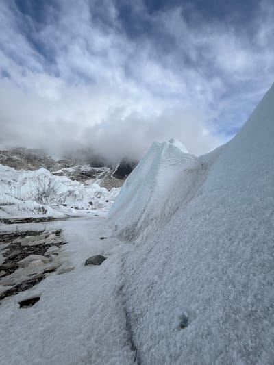 The image presents a dramatic high-altitude mountainous landscape, dominated by immense formations of snow and ice. The perspective appears to be looking up or along a steep incline, with a towering wall of glacial ice filling the right side of the frame. This ice wall exhibits a range of textures, from granular snow to smoother, denser ice with subtle blue tints, indicative of its depth and age. Darker, rough rock surfaces are visible protruding through the snow and ice, particularly towards the lower and middle sections of the image.
The left side of the frame is occupied by a expansive sky, characterized by a dynamic blend of light blue patches and soft, broken clouds in shades of white and grey. Further into the distance on the left, a rugged mountainous terrain, heavily covered in snow and ice, is partially shrouded in atmospheric haze or clouds. Jagged peaks and extensive glacial features are discernible, emphasizing the formidable nature of the environment.
No people or artificial structures are visible, highlighting the remote and untouched character of the location. The scene conveys a sense of vastness, isolation, and natural grandeur. The visible snow and ice strongly suggest cold temperatures. Given the context of Khumjung, Nepal, this landscape is consistent with the high-altitude glacial regions found in the Himalayas. The lighting indicates it is daytime, though the exact time is not discernible. No visible text is present in the media.