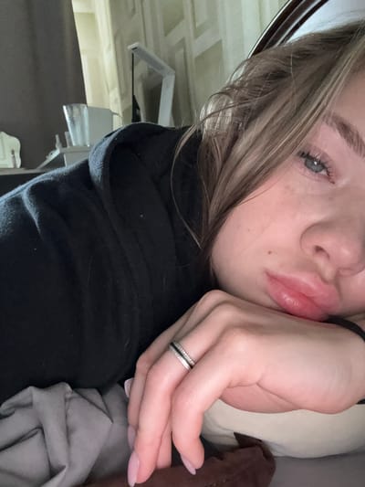 This is a close-up, low-angle selfie of a young woman. She is lying down, with her head resting on a light-colored pillow, and her left hand is supporting her face. She has blonde hair that is partially obscuring her face, blue eyes, and light skin with freckles. Her lips are full and slightly parted. She is wearing a black hooded sweatshirt. A silver ring with black and clear stones is on her left ring finger. Her fingernails are painted a pale pink. The background is blurred but appears to be indoors, with what looks like a bed or couch with gray and brown bedding. The lighting suggests it could be daytime, but the exact time of day is unclear. The woman's expression seems relaxed or possibly thoughtful. There is no visible text in the image. The location context is Severnoye, Russia.