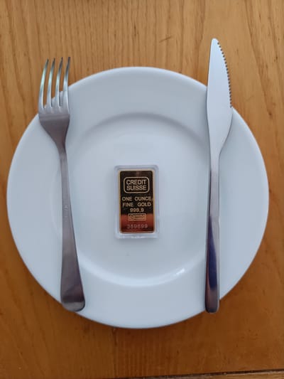 The image depicts a white plate positioned on a light brown wooden table. A fork is placed to the left of the plate, and a knife to the right. Centered on the plate is a small, rectangular gold bar enclosed in clear plastic packaging. The gold bar has text printed on it, including "CREDIT SUISSE", "ONE OUNCE", "FINE GOLD 999,9", and a serial number "389699". The scene appears to be a still life arrangement with no people or explicit actions taking place. The setting does not provide any specific indicators of its location within the city of Vitoria-Gasteiz, Spain.