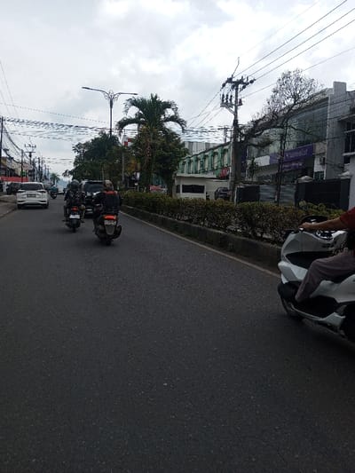 This image depicts a street scene under an overcast sky in Khajuripada, India. The asphalt road, appearing dry, features two traffic lanes moving away from the viewer, separated from the opposite direction by a raised concrete median strip topped with dark green hedges.
In the immediate foreground on the right, the front portion of a white scooter, likely a Honda PCX, is visible, operated by an individual wearing a red shirt and light-colored trousers. Ahead on the left lane, two dark motorcycles, both scooters, are in motion, heading away from the viewer. The lead motorcycle's driver wears a dark jacket with orange accents and a helmet, with license plate "B 5275 VBJ" visible. The second motorcycle's driver also wears a dark jacket and helmet, with license plate "B 6005 VME". Further back in the same lane, a white SUV is visible, followed by other vehicles.
The left side of the street is lined with commercial buildings and utility poles carrying numerous overhead power lines. A tall, ornate street light pole stands above the road in the mid-distance. The right side of the street features more buildings behind the median, including a prominent multi-story grey building with purple signage, bearing the text "Muamalat." Adjacent to it is a building with green paneling on its upper floors. A large palm tree and other assorted trees are visible throughout the scene, interspersed with buildings and utility infrastructure.