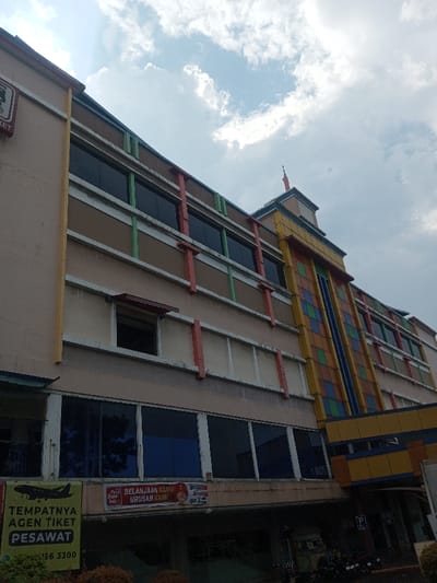 A low-angle shot shows a multi-story building with a blend of architectural styles. The building features beige walls accented with vertical stripes of red and green. Windows appear tinted, reflecting the sky. A section with a colorful, grid-like facade adds a modern touch. Visible signs include "TEMPATNYA AGEN TIKET PESAWAT." A tall structure tops the building's corner, resembling a miniature tower. The sky above is partly cloudy.