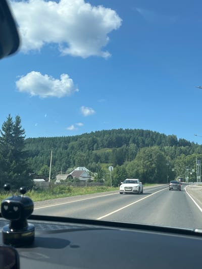 A daylight, eye-level, long shot captures a rural road in Ir'yak, Russia. A black, decorative object, resembling a robot with heart-shaped eyes, sits on the dashboard in the foreground. The road curves slightly to the right, with two lanes of traffic heading towards the distance. A white Audi sedan is in the left lane, and a dark-colored sedan is in the right lane. The landscape features a dense forest on a rolling hill in the background, with several houses visible at the base of the hill. The sky is clear blue with scattered white clouds. The scene appears to be a sunny day, typical of summer or early autumn in the region.