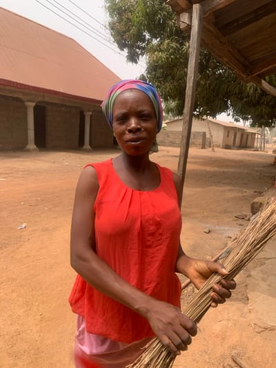 An adult Black woman stands outdoors in Garaku, Nigeria. She faces the camera directly with a neutral expression, her mouth slightly open. Her head is covered with a multi-colored headscarf. She wears a coral-red sleeveless top over a light pink garment. Her hands are clasped at waist level, holding a bundle of thin, dried sticks. The ground is unpaved reddish-brown dirt. In the background to the left, a building with a reddish-brown corrugated metal roof and unfinished concrete block walls features three white fluted columns. To the right, several smaller concrete block structures and a large tree with green foliage are visible. A wooden post and part of an overhead wooden structure are present in the right foreground. Overhead wires are visible in the upper left background against a light sky.