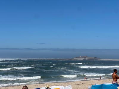 The image captures a sunny coastal scene in the municipality of Caminha, Portugal, featuring Forte da Ínsua. The upper half of the frame shows a clear blue sky with a distinct, low-lying band of atmospheric haze or distant land on the horizon. The middle ground is dominated by a dark blue-green ocean with numerous white-capped waves rolling towards the sandy shore.
In the mid-distance, slightly right of center, Forte da Ínsua, a stone fort with visible walls and structures, stands on a rocky island amidst the water. The fort appears well-preserved and is surrounded by dark water and rocky outcrops.
The foreground consists of a light brown sandy beach. On the right, a shirtless male with light skin and dark hair stands, facing left, wearing patterned swim shorts. Near the water's edge, two individuals are walking away from the viewer; one wears a reddish top and dark bottoms, the other lighter clothing. A third individual is partially visible further right at the water's edge. In the far left foreground, the top of a person's head with dark hair is visible. Various beach shelters are present in the foreground: a bright blue fabric windbreak on the right, and white fabric panels in the center, one of which has a straw hat resting on it.