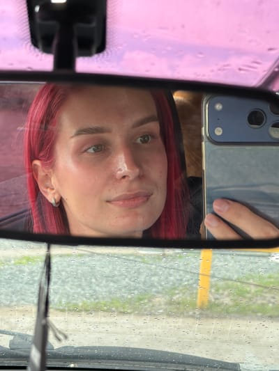 An upside-down close-up shot captures the reflection of a person in a car's rearview mirror, facing the viewer. The individual, likely a young female, has prominent red or magenta-colored hair that frames their face and partially covers their shoulder. Their eyes, a light shade of brown or green, are open and look directly towards the mirror's perspective. The person's expression is neutral, with slightly parted lips. A small, silver hoop earring is visible in their left ear.
A hand, presumably the person's, is visible holding a smartphone with a blue protective casing, which features a dual-camera setup and a flash. The phone is positioned above the person's head, partially obscuring the top of their forehead and hair in the reflection.
The background visible through the wet car windshield, outside the mirror's frame and to the right, shows numerous rain droplets, indicating recent or ongoing precipitation. Beyond the water-streaked glass, a blurry outdoor scene reveals green vegetation and a horizontal yellow line, possibly a road marking or curb, suggesting an outdoor parking lot or roadside setting. The left side of the reflection, outside the mirror, shows a pinkish or purplish hue, possibly from tinted glass or an object inside the car. The overall setting appears to be inside a vehicle during daytime in Kamyennoye, Russia, with overcast or rainy weather conditions. No discernible text is visible in the image.