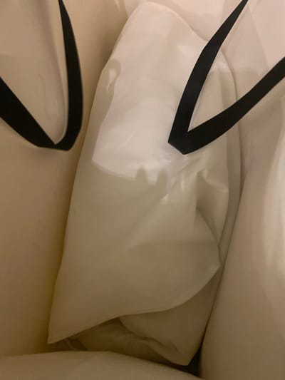 The image shows the interior of a white shopping bag, viewed from above. The bag contains several items wrapped in white tissue paper, some of which are partially visible. Black ribbon handles of the bag are also visible, one on the left and one extending from the top left corner. The lighting suggests an indoor setting, possibly at night or in a dimly lit room, as the overall tone is muted and shadows are present. The scene appears to be within Buynaksk, Russia. There is no text visible in the image.