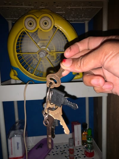 In the city of Karangkemiri, Indonesia, on a white shelf, a yellow and blue fan resembling a cartoon character is visible. A hand is holding a set of keys. One key, made of brass, is held by the ring. Two other keys, one black and one grey, are attached to the same ring. Below the shelf, a pack of tissues, a purple object, and several small bottles and containers are present. The background is a blue wall.