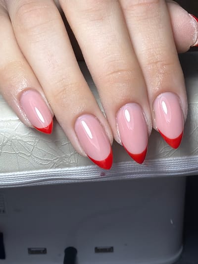 The image displays a close-up of a human left hand, featuring four fingers and part of a fifth, resting on a light-colored, textured surface. The nails are manicured with an almond-pointed oval shape, exhibiting a glossy, translucent pink base coat and sharply defined, vibrant red V-shaped tips, indicative of a contemporary French manicure. Slight white particulate residue is present on the skin around the nail beds. The hand rests upon a light, possibly off-white, ribbed or patterned surface, which appears to be the top of a nail curing lamp, with a darker, blurred background below. This scene is situated in the city of Kavrakirovo, Bulgaria.
