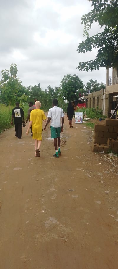 A group of people are walking away from the camera on a dirt road in Achara Ndiagu Omegio, Nigeria. The weather appears overcast, with a cloudy sky.
In the foreground, a person with albinism, wearing a bright yellow t-shirt and shorts, walks with another person in a white short-sleeved shirt and green shorts. Behind them, a person in a black t-shirt and dark pants is visible, along with a partially obscured figure in a dark shirt. Further ahead, a woman in a black dress walks away from the camera.
To the right of the road, a partially constructed building is visible, with stacks of bricks and building materials nearby. A sign with text is placed in front of a small structure. The area is surrounded by lush green vegetation and trees, suggesting a rural or semi-rural setting. The road is muddy and uneven, with some puddles, indicating recent rain. The overall mood seems casual, with individuals walking together. No specific event or emotional expression is evident.