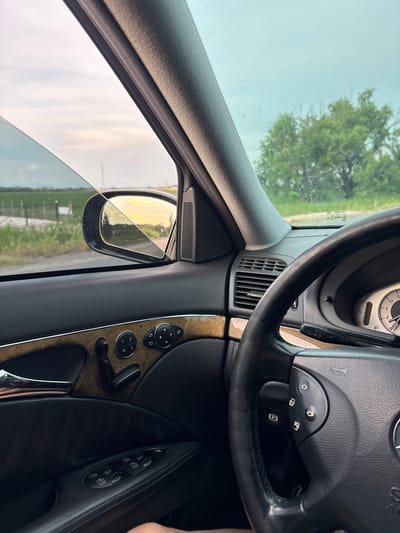 The image is a first-person perspective from inside a car, looking out the driver's side window and towards the steering wheel. The car appears to be parked on the side of a rural road in Yoakim Gruevo, Bulgaria. The interior of the car shows black leather upholstery on the door panel, wood trim accents, and a steering wheel with various controls. The window is slightly down, revealing a hazy sky with soft, warm light suggesting either sunrise or sunset, casting a golden reflection in the side-view mirror. Outside, there's a green landscape with fields and trees, bordered by a fence and a road. The overall impression is one of a peaceful, rural setting.