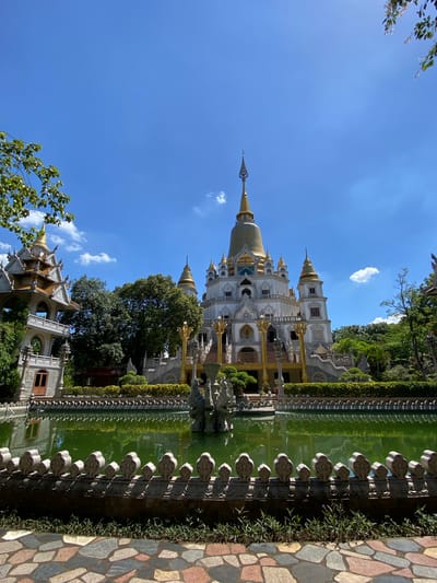 The image depicts a temple complex situated in the city of Dĩ An, Vietnam. The main structure is a tall, multi-tiered building with a golden dome and white facades, resembling a Buddhist pagoda or stupa. It is flanked by other ornate buildings with similar architectural styles. The complex is surrounded by lush greenery, including trees, shrubs, and manicured hedges. A still, green body of water reflects the temple's image. A decorative stone fence lines the edge of the water feature, with intricate patterns. The sky is blue with scattered clouds, suggesting a bright, sunny day. The perspective is from a low angle, emphasizing the height of the temple.