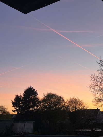 The image shows a sky at dusk in Würselen, Germany. The sky has a gradient of colors, transitioning from a pale blue-purple at the top left to a peachy orange towards the horizon line. Several contrails cut through the sky. Silhouetted trees and building edges frame the view, obscuring details of the surrounding environment.