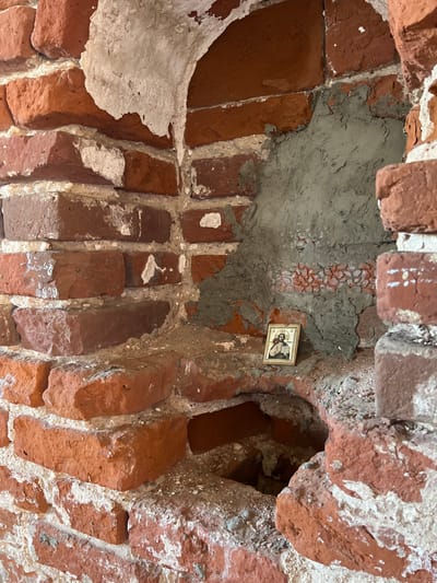 The image shows a section of an old brick wall and archway. The bricks are red and partially covered with white plaster or mortar. A patch of gray mortar fills a section near the top. A small religious icon is affixed to the gray mortar. The location is Шуран, Russia.