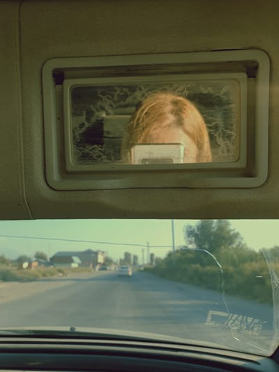 The image shows a reflection in a car's sun visor mirror of a person's face, possibly a woman with blonde hair, holding a smartphone. The background of the image is a blurred view of a road, with buildings and trees visible on the side. The scene appears to be set in Griboyedov, Armenia.