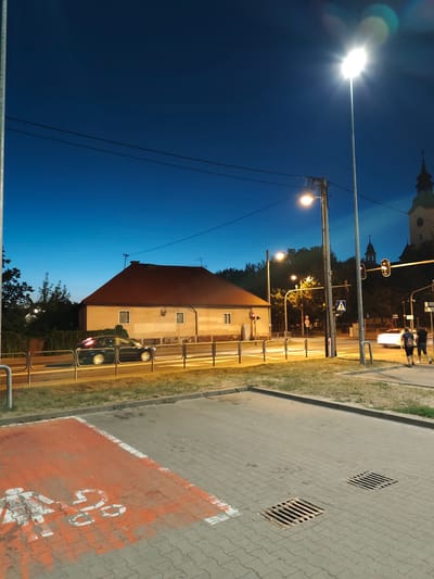 A low-angle, nighttime, exterior, full shot captures a street scene in Stryków, Poland. The sky is a deep, clear blue with a bright streetlight dominating the upper right portion of the frame. Power lines crisscross the sky. In the middle ground, a two-story, light-colored building with a red-tiled roof is visible. A dark-colored car is driving past the building, its headlights and taillights blurred due to motion. A metal fence runs along the side of the road in front of the building. To the right of the building, a traffic light displays red. Further to the right, two individuals, one with dark hair and a backpack and another with lighter hair, are walking on the sidewalk. In the foreground, a paved area with cobblestones is visible, with two metal grates embedded in the pavement. An area marked with red paint and white symbols, possibly indicating a parking space or a designated area, is also present in the foreground. The overall impression is a quiet, urban street at dusk or early night.