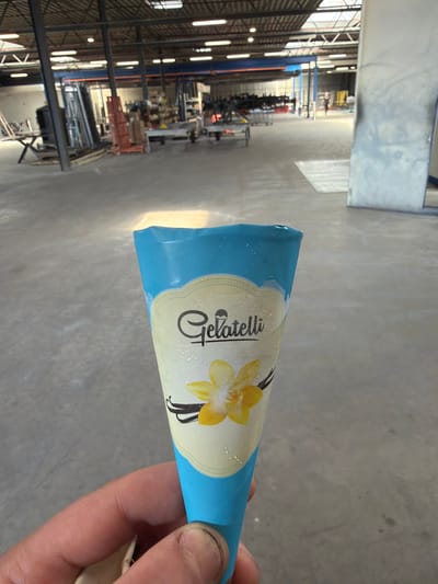 The image shows a hand holding a vanilla ice cream cone. The cone is wrapped in blue and yellow packaging with the brand name "Gellati" clearly visible in a stylized font. A yellow vanilla flower and two vanilla pods are depicted on the packaging. The cone appears to be slightly melting, with small droplets of condensation on the wrapper.
The background is a blurry industrial setting, likely a warehouse or factory, with metal shelving, lighting fixtures, and various objects stored on the shelves. The floor is a concrete surface. The lighting suggests it is daytime. The image appears to be taken in Mol, Belgium, as indicated by the location context. The hand holding the cone is the only visible person in the image. The overall impression is casual and informal, possibly indicating a break or a moment of personal enjoyment.