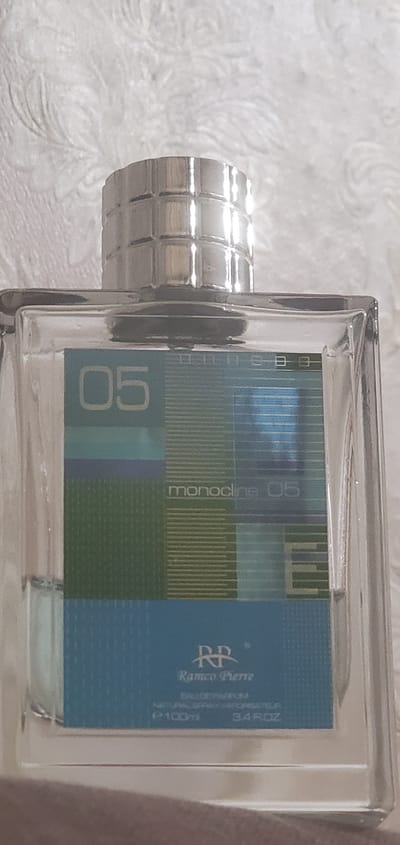 A close-up, angled shot shows the top portion of a clear glass perfume bottle. The bottle has a silver, ribbed cap. The label on the bottle is a design of horizontal lines in varying shades of blue, green, and light blue. The number "05" is prominently displayed in a light blue rectangle on the left side of the label. Below this, the word "monocline" appears in white text, followed by "05". Further down, the logo "RR" is visible above the text "Ramco Pierre". Below this, "EAU DE PARFUM" and "NATURAL SPRAY VAPORISATEUR" are printed in smaller text, along with "100ml" and "3.4 ROZ". The background is a textured, white surface with a swirling pattern, likely fabric. The scene is not in Konakovo, Russia, as there is no identifiable urban or geographical context.