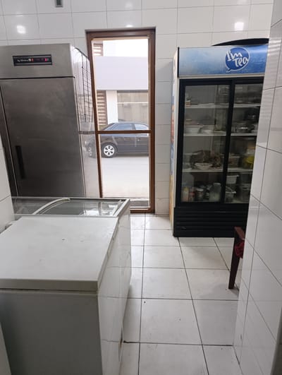 The image shows an interior scene, likely a commercial kitchen or food service establishment in Buzovna, Azerbaijan. In the foreground, a white chest freezer is partially visible. To the right of the freezer, a large, glass-fronted refrigerator with a blue and white "I'm Tea" logo is prominently displayed, filled with various food items and containers. Behind these appliances, a wide doorway with a wooden frame opens to an outdoor area where a dark-colored sedan is parked. To the left of the doorway, a tall, stainless steel commercial refrigerator is positioned. The floor is tiled with white squares, and the walls are covered in white tiles. The scene appears to be a functional workspace, with appliances ready for use.