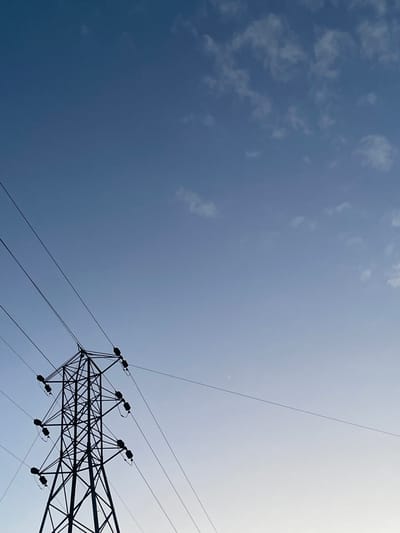 This low-angle shot captures a silhouetted electrical pylon against a vast, clear sky with a few wispy clouds. The pylon, a latticework of dark metal, dominates the right side of the frame, its intricate structure supporting multiple power lines that stretch across the sky. The sky transitions from a deep blue on the left to a lighter, brighter shade towards the right, suggesting the sun is positioned in that direction, possibly setting or rising. The overall scene evokes a sense of open space and the industrial infrastructure that hums beneath the natural elements. The context of Kassa Market, Nigeria, places this scene within a specific geographic and economic landscape, hinting at the vital role of power transmission in the region. The weather appears clear and pleasant, with no indication of rain or extreme conditions. The image is devoid of any people or visible text.