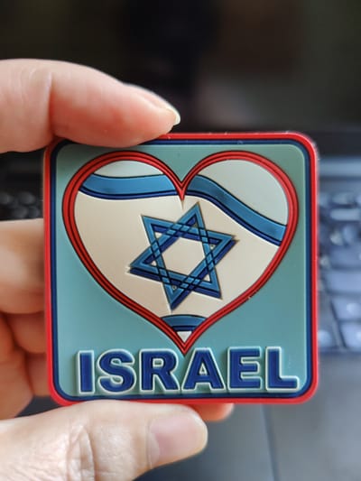 A square magnet with a red border is held by a person's fingers. The magnet features a heart shape with the Israeli flag inside, a blue Star of David in the center, and the word "ISRAEL" in blue letters at the bottom. The background appears to be a dark surface, possibly a laptop keyboard. The image does not show any location in the city of Rybnitsa, Moldova.