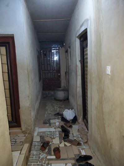 This image depicts a narrow, dimly lit hallway in a residential building located in Ikorodu, Nigeria. The hallway has textured, light-colored walls and a low, corrugated metal ceiling. The floor is a mosaic of small, irregular tiles in various shades of cream, gray, and brown.
Several pairs of footwear are scattered across the floor, suggesting recent activity or storage. These include dark-colored sandals and flip-flops, as well as a pair of brown leather sandals. A white plastic bag is also visible on the floor.
Further down the hallway, a large, metal cooking pot sits near a metal gate that appears to be an entrance to another room or area. This gate is secured with a padlock. Adjacent to the pot is a doorway with a dark-colored frame and a window with metal bars. On the right wall of the hallway, a white light switch is mounted. The overall scene suggests a functional, perhaps utilitarian, living space within a Nigerian urban environment.