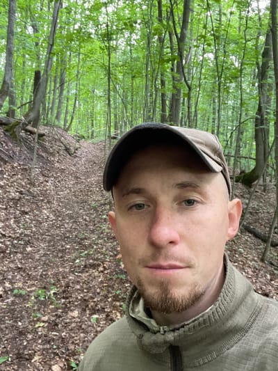 A man wearing a brown baseball cap and a green jacket is taking a selfie in a forest. The man has short brown hair, a light complexion, and a goatee. He is looking directly at the camera. The background is a dense forest with tall trees and fallen leaves on the ground. There is a path winding through the trees. The location is the city of Nikolayevka, Russia.