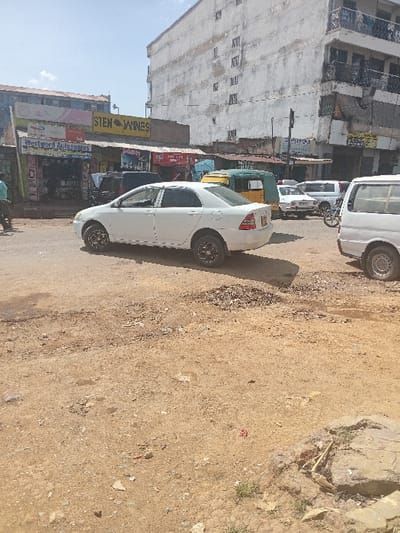 The image, rotated counter-clockwise, depicts an outdoor scene, likely a street or unpaved road in an urban or semi-urban area, under clear daylight conditions. The ground is a light brown, dusty, and uneven surface, appearing to be dirt or compacted earth rather than paved asphalt, with some scattered debris and small rocks visible, particularly on the right side.
In the mid-ground, several vehicles are visible. A white sedan, possibly a Toyota, is parked or driving on the left side of the dirt road, facing away from the viewer towards the background. It has dark-colored alloy wheels. Behind it, a green and yellow auto-rickshaw (tuk-tuk) is partially visible. Further back, several other vehicles, including at least one white van and another white car, are seen on what appears to be a paved or more formal road section.
On the left side of the image, a row of buildings lines the street. The most prominent building on the far left is a multi-story structure with a light, possibly off-white or faded grey, facade. It shows signs of wear and tear, with some discoloration and apparent age. Multiple small, rectangular windows are visible across its face.
Below this large building, and extending towards the foreground, are smaller commercial structures. Several shopfronts are visible with various signs. One prominent yellow sign reads "STEN WINES" in bold, dark letters, suggesting a liquor store. Below it, another sign, partially obscured, appears to read "Westwood Autospares," indicating an auto parts store. Other signs with colorful graphics and text, mostly in an unidentifiable foreign language (likely an Indian script given the auto-rickshaw), are also present but are too small or blurry to be fully deciphered.
The sky is a bright, clear blue, visible in the upper left corner, suggesting fair weather. The overall impression is that of a bustling street scene in a developing country, characterized by a mix of residential/commercial buildings, diverse vehicles, and unpaved ground.