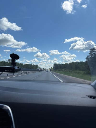 The image presents a sideways view from inside a vehicle, looking out onto a highway under a bright sky. The perspective is from the front passenger or driver's side, through the windshield.
In the immediate foreground, to the right when viewed upright, is a portion of the vehicle's interior, likely the dashboard or A-pillar, and a dark-colored device with a suction cup mount, possibly a dashcam or phone holder, attached to the windshield.
The central part of the image features a multi-lane highway or road stretching into the distance. A clear white line marks the lane separation. A metal guardrail runs alongside the road, beyond which is a dense, lush green forest that extends along the entire visible stretch of the road. Several other vehicles, including cars and trucks, are visible in the far distance on the highway, indicating active traffic.
The background is dominated by a vibrant blue sky dotted with numerous fluffy, white cumulus clouds, suggesting fair weather.
The scene depicts an activity of driving or traveling along a highway. Based on the bright illumination and clear sky, it is daytime, likely midday or early afternoon, with sunny conditions and scattered clouds. The dense forest alongside the road suggests a rural or semi-rural setting.
The location context provided for this image is Poddubki, Russia. No discernible text is visible within the frame.