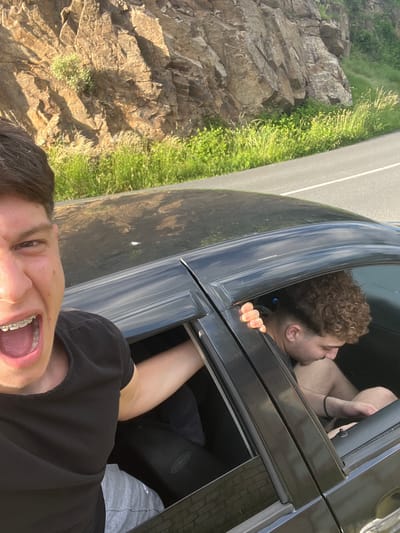 This is a medium shot, eye-level selfie taken outdoors with a car and a rocky, grassy hillside in the background. In the foreground on the left, a young male with dark hair is looking directly at the camera with his mouth open and braces visible. He is wearing a black t-shirt. His left arm is extended out of the car window. Inside the car, on the right side, another young male with curly brown hair is looking down. His right hand is resting on the car door frame. He is wearing a dark t-shirt and shorts. The car is dark colored and the windows are open. The background consists of a steep, textured rock face with green grass and vegetation growing at its base and along a road. The road appears to be paved and has a white line marking. The lighting suggests it is daytime, likely with sunlight illuminating the scene. There is no specific indication of the city of Pechinska, Bulgaria, within the image itself.