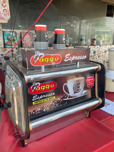 Here is a detailed description of the image:

**Scene Overview:**

The image shows a mobile espresso coffee machine situated on a red surface, likely a vendor's stand. The background reveals a view through a glass partition, displaying items on shelves, bottles, and religious pictures. The scene appears to be in a location with a mix of commercial activity and possibly a residential area.

**Objects:**

*   **Espresso Machine:** The coffee machine is the main focus, with prominently displayed "Paggo Espresso" branding and a design that includes chrome bars, a coffee cup graphic, and coffee bean imagery.

*   **Coffee Cups:** A stack of black coffee cups with red and white stripes is placed adjacent to the espresso machine.

*   **Shelves:** In the background, shelves contain bottles, containers, and various products.

*   **Religious Pictures:** Beneath the coffee cups, there are framed religious pictures.

*   **Advertising materials:** Some type of advertising or promotional materials are visible on the left and upper portion of the photo.

**Environment:**

The scene seems to be outdoors or in a semi-open space where street vending is likely occurring. The environment is relatively crowded or has many objects visible within a small area. The location is likely in Katauhar Kalan, India, based on the location data.