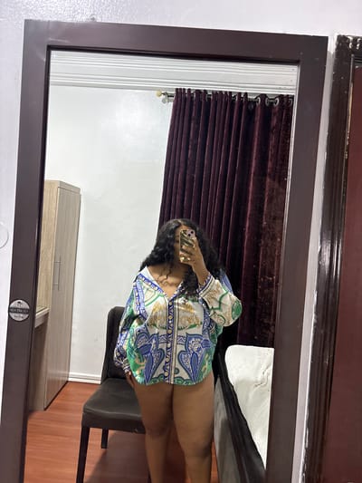 A woman stands in front of a mirror, taking a selfie with her phone. She wears a patterned short-sleeved shirt. Behind her, a maroon curtain partially covers a window. A chair is visible, and a cabinet stands against the wall. The floor is wooden. The scene suggests an interior space, possibly in a residential area, and considering the prompt's location details, presumably within Port Harcourt, Nigeria.