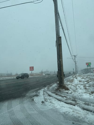 A roadside scene shows snow falling. A gray sky provides diffused light. A multi-lane road is partially covered in snow, with tire tracks visible. On the shoulder is a build-up of snow. There is a black car traveling on the road. Utility poles and wires are present on the roadside. A sign is also visible, along with what seems to be a gas station in the distance.