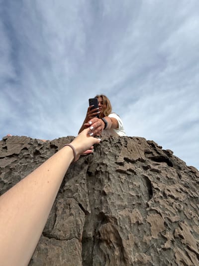 A low-angle shot shows a person extending their arm and hand towards another person positioned at the edge of a rocky precipice. The person at the edge is holding a smartphone, seemingly taking a photo, and is reaching down with their other hand towards the extended hand. The location is identifiable as the city of Seoca in Montenegro, based on the unique geological features and rocky landscape. The sky with scattered clouds is visible in the background.