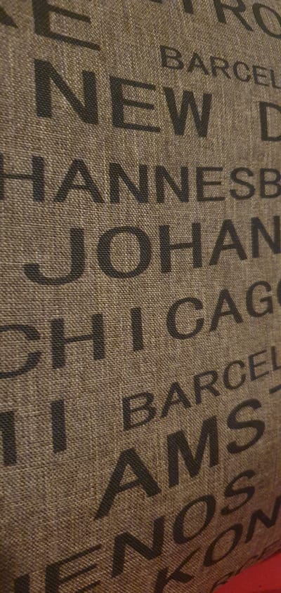 The image displays a close-up, textured view of fabric with large, dark, sans-serif lettering. The text appears to be a list of city names, vertically oriented and running in columns. Visible cities include "NEW D", "HANNE SB", "JOHAN", "CHICAG", "BARCEL", "AMS", and "NOS N". The fabric has a woven texture, appearing to be a coarse, burlap-like material in a muted brown or tan color. The lighting creates subtle shadows, highlighting the texture of the fabric and the raised surface of the letters. The bottom right corner of the image shows a sliver of a bright red surface, possibly another object or a background element, which is out of focus. No specific location in Alūksne, Latvia, can be identified from this image, as it focuses solely on the pattern of the fabric.