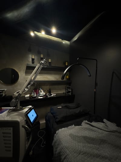 The scene is an interior space, possibly a medical or cosmetic treatment room. The location is Ciudad López Mateos, Mexico.
The room has a dark, contemporary aesthetic with dark gray or black walls. There are vertical shelving units along the wall, filled with various bottles, containers, and decorative elements, including small potted plants and cylindrical items resembling candles.
A specialized medical device, likely a laser or similar treatment machine, is prominently positioned in the foreground. It features a touch screen interface displaying a blue screen with a white play button icon. The device has an articulated arm extending towards the shelving unit. The text "Chrome" is visible on the device's side panel.
A circular mirror with a gold frame is located below the shelving units and the arm of the medical device.
Lighting is provided by recessed lights in the ceiling and an arc-shaped lamp stand.
To the right, a bed or treatment table is partially visible, draped with a gray textured blanket. A dark-colored garment or bag is also present on the bed.
