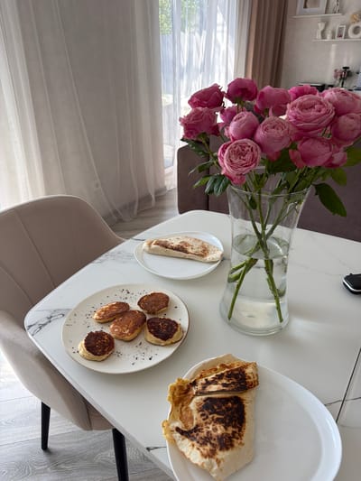 On a white marble-patterned table in Malinovka, Russia, a bouquet of pink roses in a clear glass vase is centrally positioned. To the left of the roses, a white plate holds several small, round, golden-brown pancakes, sprinkled with dark seasoning. Adjacent to this plate, another white plate contains a folded flatbread with char marks. In the foreground, a white oval plate displays a folded flatbread, partially grilled. A beige upholstered chair with black legs is visible to the left of the table. Behind the table, sheer white curtains are drawn across a window, allowing sunlight to stream in. The background shows a glimpse of a sofa and shelves with decorative items.