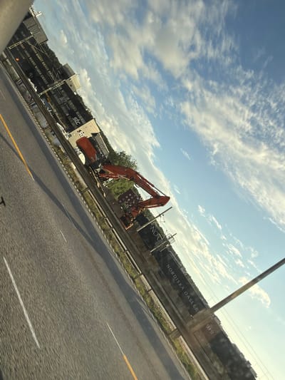 An orange excavator is parked on the side of a highway in Mölndal, Sweden. The highway appears to be under construction, with barriers and construction materials visible. There are also shipping containers in the background, some of which have the "STC" logo on them. A sign that reads "NORDISKA GALLER" is also visible. The sky is blue with some white clouds. The image is taken from a moving vehicle, as evidenced by the motion blur.