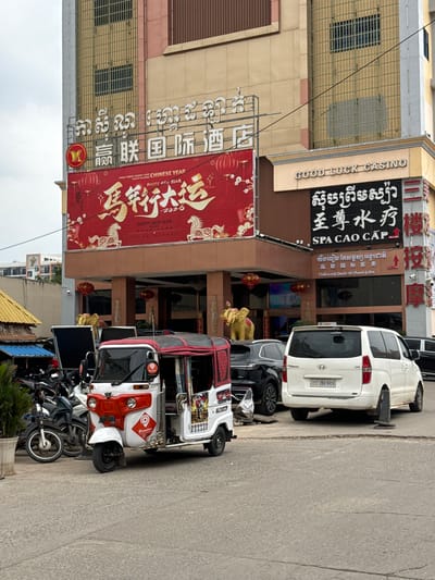The image depicts a street scene in the city of Bavet Quarter, Cambodia. On the left, there's a tall building with multiple establishments, including "Good Luck Casino" and "SPA CAO CAP" (a spa). Chinese characters and banners promoting the Chinese New Year 2026 are also visible. Vehicles, including cars and tuk-tuks, are parked along the side of the road. Several motorbikes are parked together near the bottom of the frame. The sky is overcast, suggesting a dull day. The overall environment appears to be a busy street, likely a commercial area with shops and services.