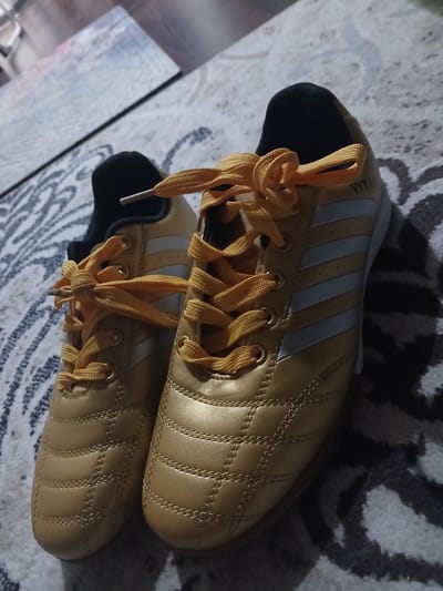 A pair of gold soccer cleats with yellow laces is displayed on a rug. The cleats have white stripes on the sides and the word "VITAL" is visible on the right cleat. The background consists of a rug with a black and white patterned design and a blurry image of what appears to be a wall or a picture. The lighting suggests an indoor environment, possibly a home. There is no information to determine the location in the city of Tuchkovo, Russia.
