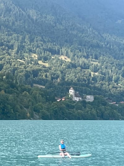 The image depicts a daytime scene on Lake Brienz in Bönigen, Switzerland. In the foreground, a single individual is seated on a light-colored stand-up paddleboard on the turquoise water. The individual wears a light-colored top, dark shorts, a blue life vest, and a white visor, with their legs extended forward on the board. A dark-colored paddle rests horizontally across the front of the paddleboard, alongside a dark bag or object. The background features a steep mountain covered in dense green forest, rising from the lake's edge. Numerous lighter green clearings are visible on the mountainside. Prominently situated on a forested knoll at the base of the mountain is a complex of buildings, the most distinct being a light-colored structure with a dark-roofed steeple and arched openings, identifiable as the Old Church of Bönigen. Additional traditional-style buildings with dark roofs and light walls are visible near the church and sparsely scattered higher on the mountain.