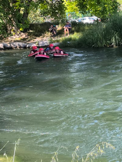 Four people wearing red helmets and life vests are in a river. They are each floating on a red and black tube-like object. Two people are on the riverbank watching them. A man sits in a chair near the edge of the river. The background has trees, grass, and a fence. The location is Saint-Pierre-de-Bœuf, France.