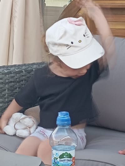 A young child wearing a white cap with a pink bow and Hello Kitty design, and a black t-shirt, is sitting on a grey outdoor sofa. The child is holding a white stuffed animal and has a bottle of water next to them. The background shows a wooden structure and a tan curtain. The scene appears to be in a backyard or patio area.
