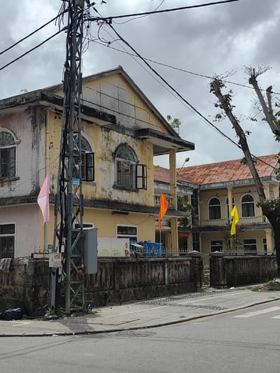 The image presents an urban street scene in Phú Xuân Ward, Vietnam, under an overcast sky. In the foreground, an asphalt road leads to a concrete sidewalk bordered by a curb. A black refuse bag is positioned on the sidewalk to the far left. A prominent, gray metal lattice utility pole, supporting numerous black electrical and communication cables that crisscross the upper portion of the frame, stands in the left-center. A gray electrical enclosure is attached to this pole.

Behind the sidewalk and utility pole, a dark, weathered stone or concrete fence with decorative ironwork encloses two multi-story buildings. The structure to the left, which is closer, features a faded light yellow and pale pink facade, exhibiting widespread staining and organic growth. It has a gabled roof section and an overhang supported by columns. Two upper-story windows with arched tops and dark-framed sashes are open. A light pink flag is affixed to a pole near the left side of this building, and an orange flag is flown further along the fence. A partially visible sign displaying "TAEKWONDO" and a martial artist graphic is mounted on the fence. Blue tarpaulins are visible behind the fence line.

Further into the scene, a second building with similar architectural characteristics, including arched windows and a yellow facade, is visible. This building is topped with a reddish-brown corrugated metal roof. A yellow flag is flown near this structure. A street light fixture is positioned between the two buildings above the fence line. To the right, several deciduous trees with sparse new leaf growth stand against the sky. No individuals are present in the depicted scene.